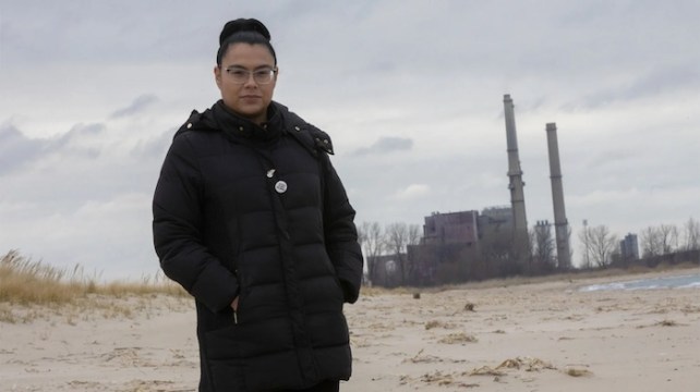 Dulce Ortiz, a woman with light brown skin, standing at a beach with a factory in the background