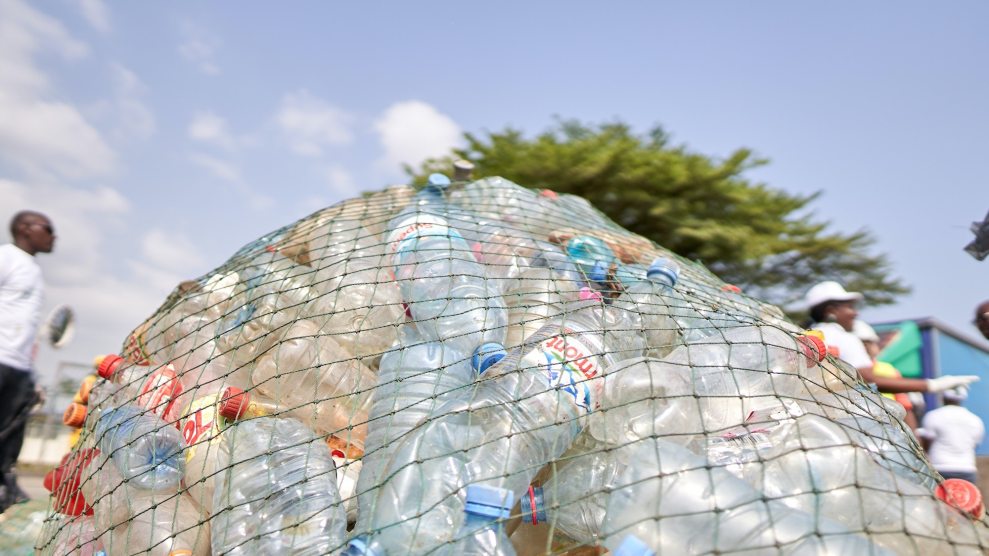 A photo of plastic bottles in a mesh bag. Black people talking can be seen in the background, and it is a sunny day.
