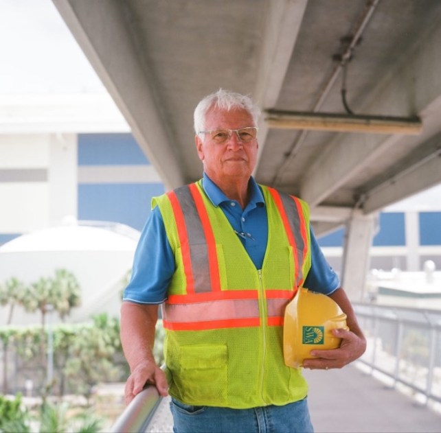 A white aging man under a highway with white hair wearing a yellow vest