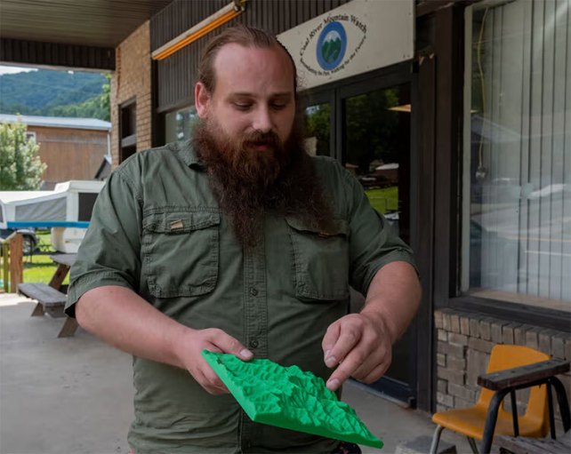 A white man with a dark copper beard and a green button down shirt points out a spot on a 3D printed model of section of West Virginia.
