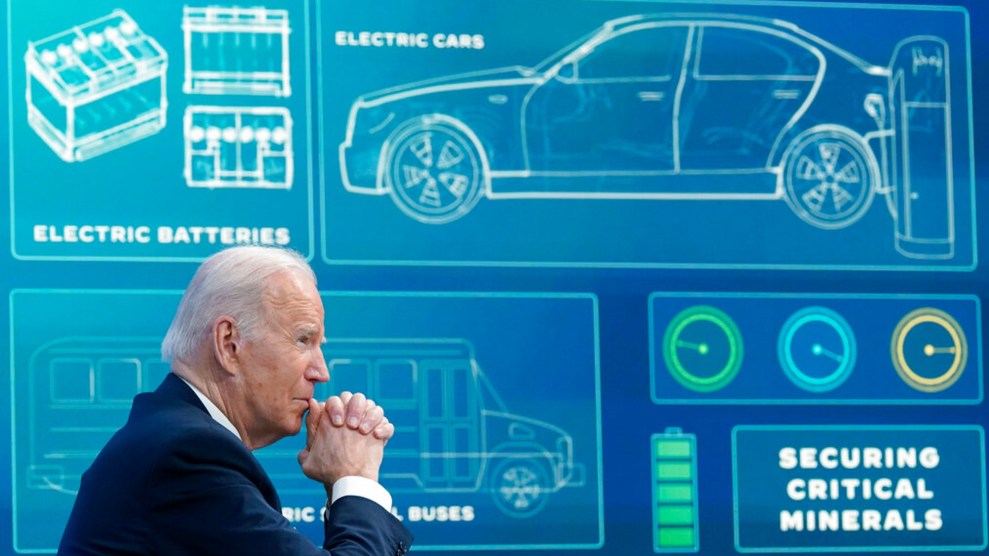Joe Biden sits with his chin in clasped hands in front of a blue board projecting information about electric vehicles, including an outline of a car and battery and the words "Securing Critical Minerals."