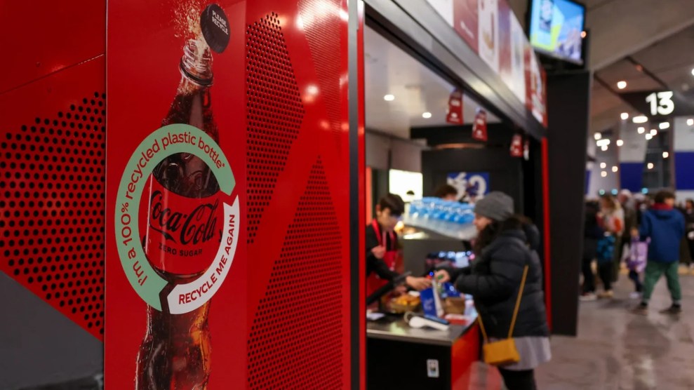 A red sign that reads "I'm a 100% recycled plastic bottle recycle me again" dominates the view of a blurry food booth where an individual in black puffy coat purchases a bag of food.