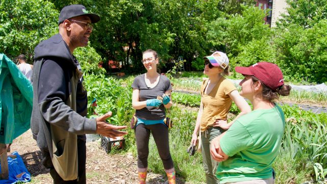 A black man stands in front of a group of white women with his arms spread open, explaining a component of the farm they all stand in.
