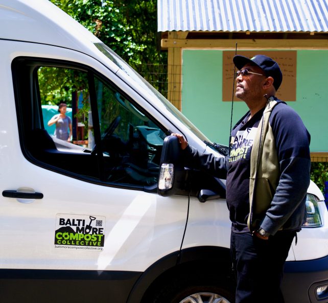 A black man with a baseball cap and sunglasses stands in front of his white, electric van.