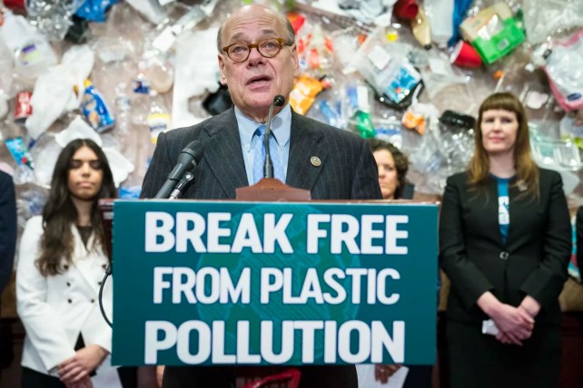A white man wearing glasses and a blue tie stands at a podium with his mouth open. Behind him, several other individuals stand with serious faces. On the wall, there is a display of plastic trash. 