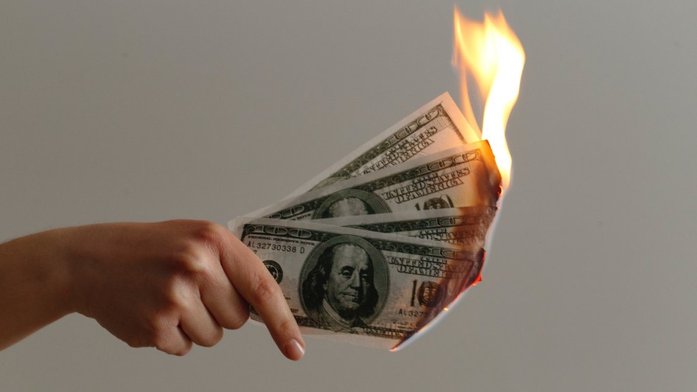 A hand that has a light skin tone holding onto hundred dollar bills that are on fire