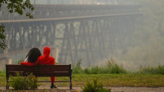A couple sits on a park bench looking at a hazy bridge.