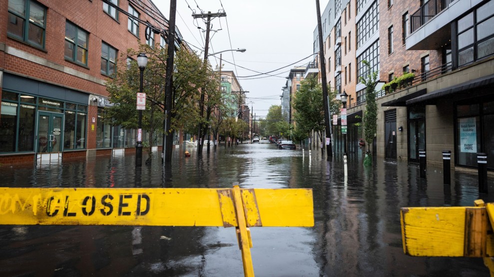 A yellow closed barrier stands in front of a flooded city street, lined with brick buildings.