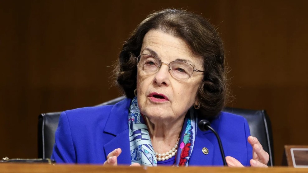 Dianne Feinstein is pictured wearing a purple-blue jacket and a pink and blue scarf. Her hair is brown and she holds her hands up, mouth open, speaking.