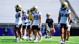 Six Washington Huskies football players at a practice in white and purple shirts and gold helmets, at their stadium. There is someone walking around with water jugs.