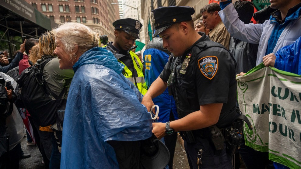 Een vrouw met wit haar en een blauwe regenjas wordt gearresteerd door een NYPD-officier met meer demonstranten en politie op de achtergrond.