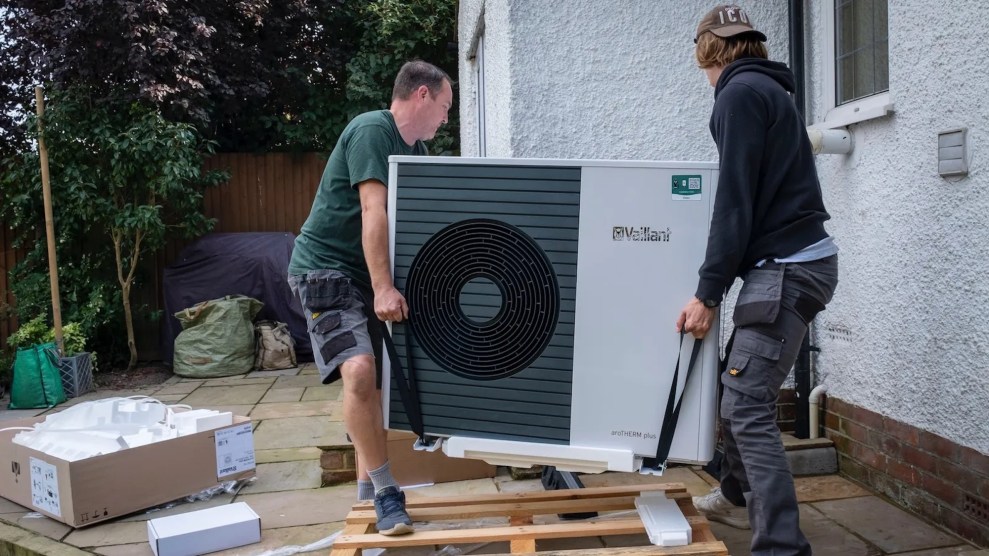 Two white people carrying a heat pump from outside to inside a home
