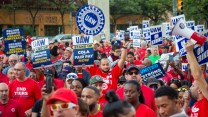 Striking UAW members in red shirts holding signs that say "UAW," "UAW: COLA Fair Pay Now" and "UAW on Strike"