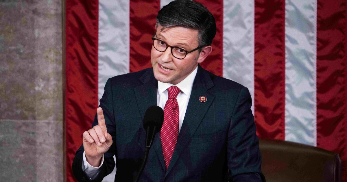Mike Johnson is wearing glasses, a dark suit with a checkered pattern, a white shirt, and a bright red tie. He stands in front of the American flag, in front of a microphone, with one finger raised as if making a point or emphasizing something during his speech. He looks earnestly towards his audience, his fellow House members, from the elevated rostrum of the House of Representatives.