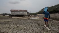 A man, holding a jug of water, walks on cracked land towards a houseboat that sits on dirt, everything is grey and brown.