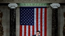 Speaker Mike Johnson standing at a podium in front of a large American flag and under an inscription "In God We Trust"