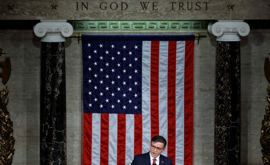 演讲者迈克·约翰逊站在讲台上，面前是一面巨大的美国国旗，下方刻有铭文 "我们信赖上帝"