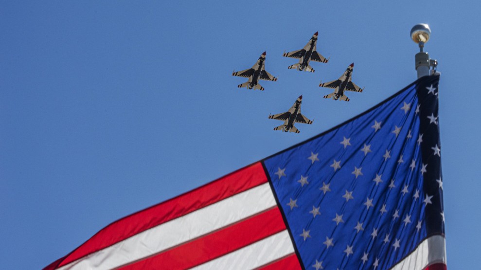 A united states flag splits a blue sky. In the remaining corner, four small black jets fly by.