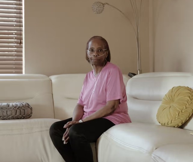 A Black woman in a pink shirt and black pants sitting on a couch with an oxygen device in her nose.