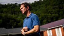 Andy Beshear, a white man with brown hair, standing outside in a blue polo with a somber expression on his face.