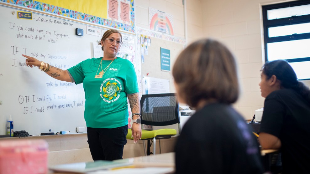 A teacher in a green T-shirt pointing to a whiteboard in a classroom.