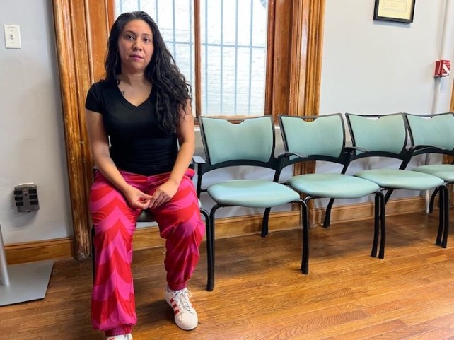 Micaela E. Martinez, a woman with light brown skin in a black shirt and pink pants, sitting on a chair