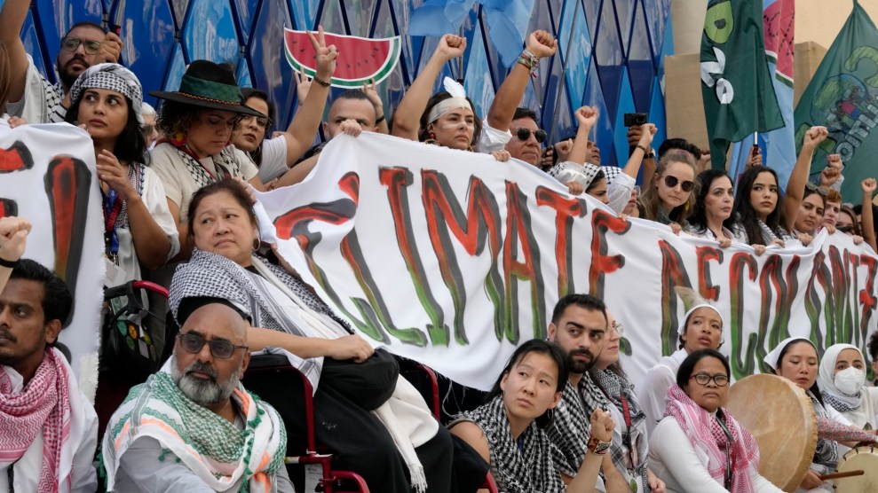 A gathering of people hold a banner that says "climate decolonize" in red and green lettering. Many raise their fists into the air and wear keffiyehs.