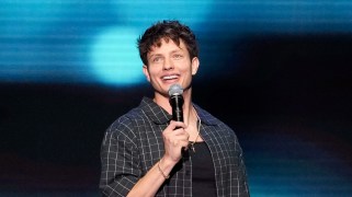 Matt Rife, wearing a plaid shirt, tank top and chain necklace, stands against a blue and black backdrop holding a microphone and smiling.
