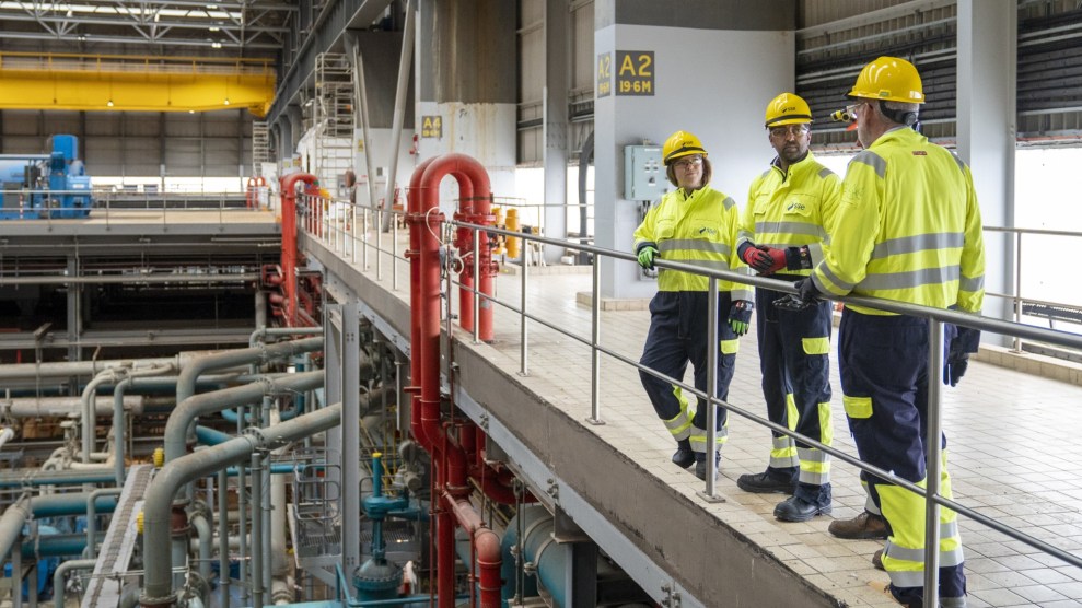 Three individuals in bright yellow vests stand on a walkway overlooking an energy plant comprised of colorful metal tubing.