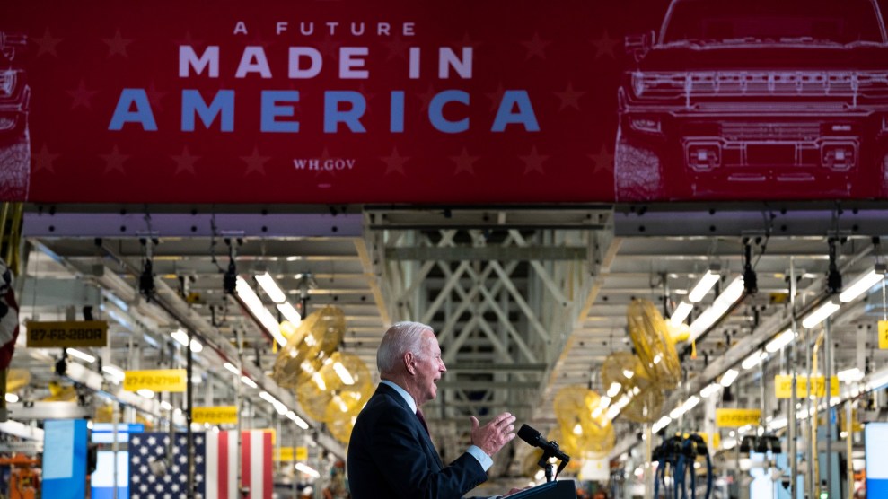 Biden stands next to a large factory floor, alit with beams and American flags. Above him a sign reading "A Future Made In America."