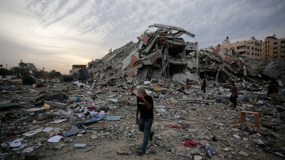 A man stands hunched in front of a scene of destroyed buildings. The sky is cloudy.