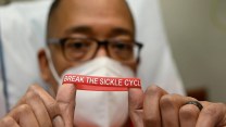 A Black man wearing glasses and a white N95 mask sitting in a hospital bed holding a red bracelet that says "Break the Sickle Cycle"