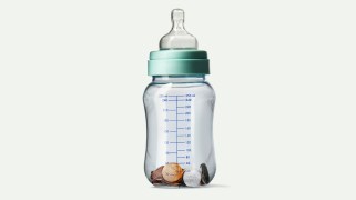 A clear baby bottle is pictured on an off-white background. Inside the bottle are a few coins: pennies, nickels, and dimes; the bottle is otherwise empty.