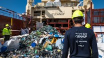 An individual in a yellow hard hat and blue shirt with "The Ocean Cleanup" faces a pile of trash on the deck of a ship.