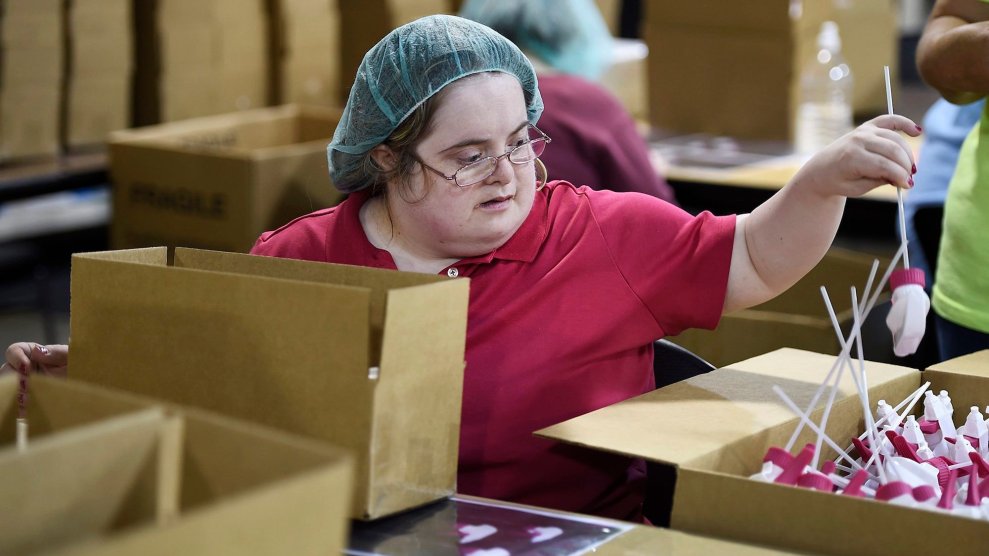 A white woman with a disability in a red shirt and hair net picking up part of a bottle spray in a cardboard box