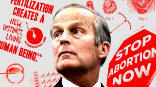 Former United States Representative Todd Akin stands in front of a gray and red collage of anti-abortion signs and various contraceptives.