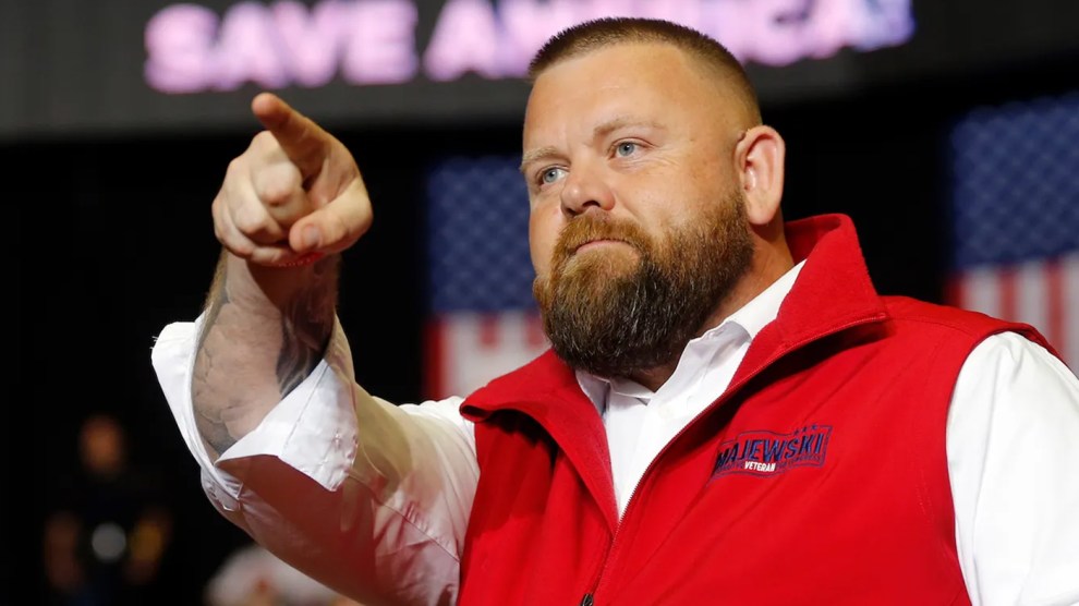 J.R. Majewski, a heavier white man with a beard, in a white shirt and red vest pointing to something off stage with a crowd surrounding him and a sign saying "Save America" in the background