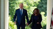 A white man wearing a blue tie walks next to a Black woman wearing a black pantsuit. They hold hands. Behind them, there is greenery and white pillars.