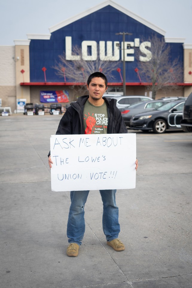 Portrait of a man holding a sign that reads, "Ask me about the Lowe's Union vote!" in a Lowes parking lot.