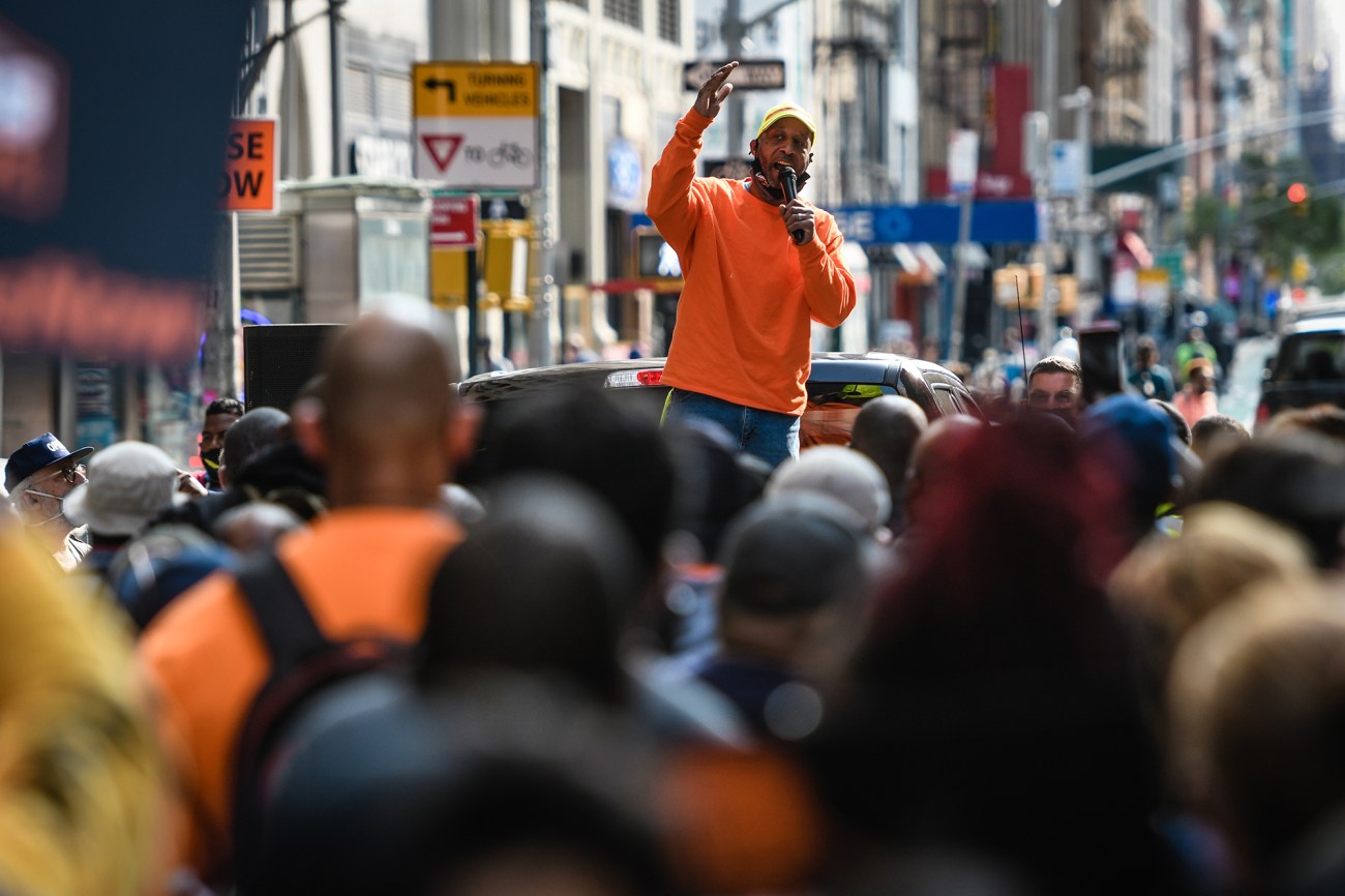 Man in orange shirt speaking to a crowd of people on the street.