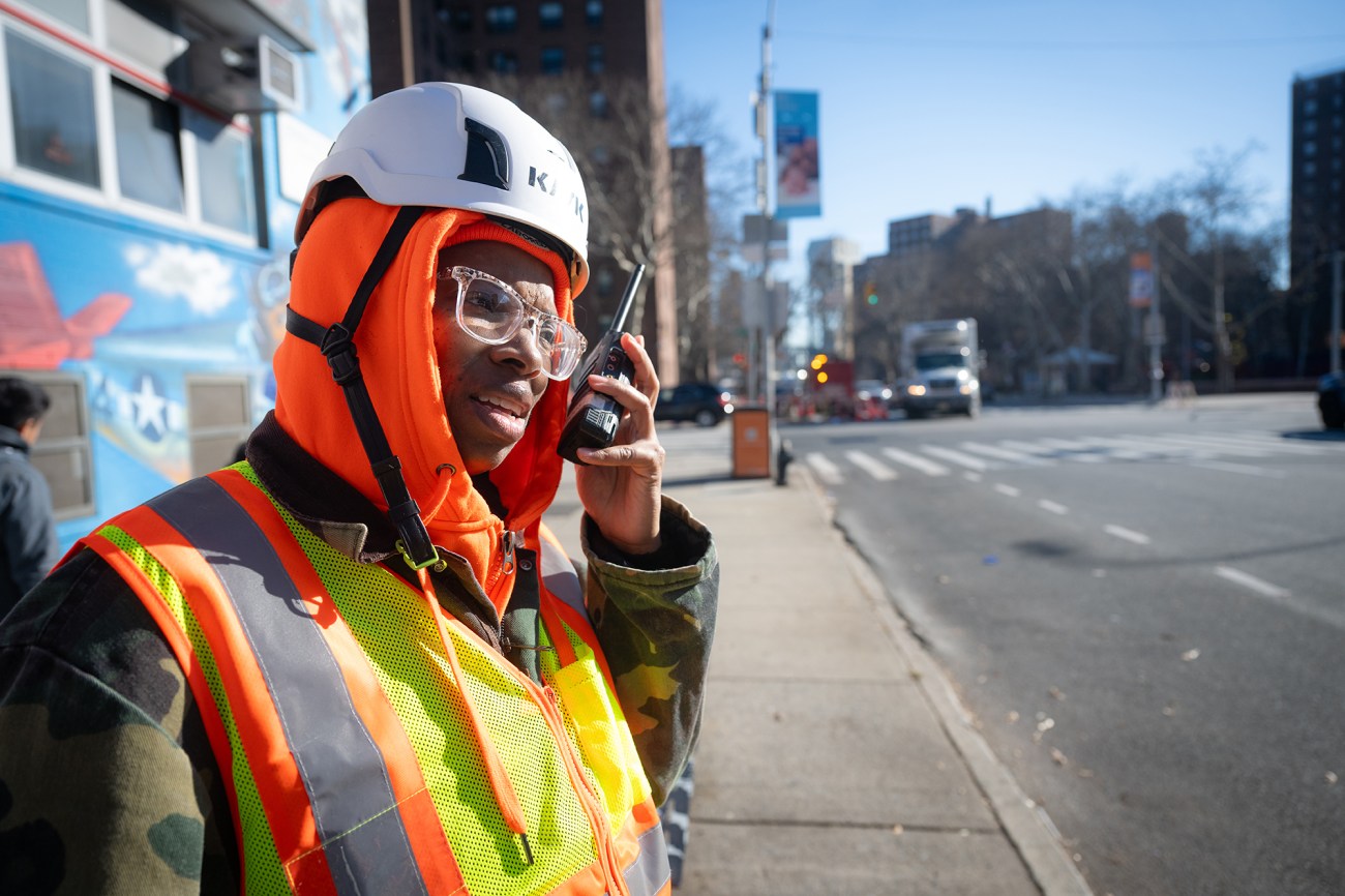 A woman wearing a yellow vest and hard hat listens to a walkie-talkie.