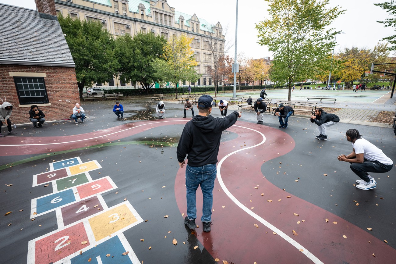 view of the back of a man standing, pointing at group of people squatting on a playground.