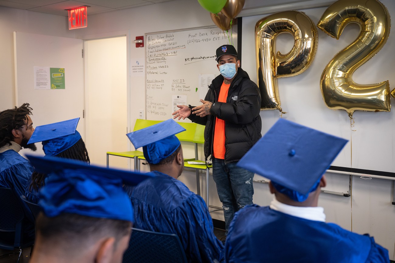 A man wearing a mask stands in front of a classroom full of students wearing blue graduating gowns.