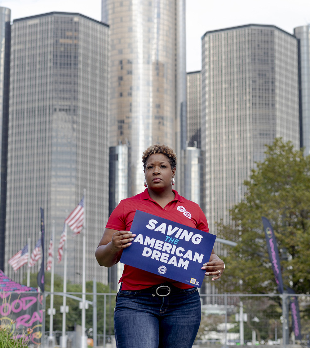 Woman standing in front of skyscraper holding a UAW protest sign.