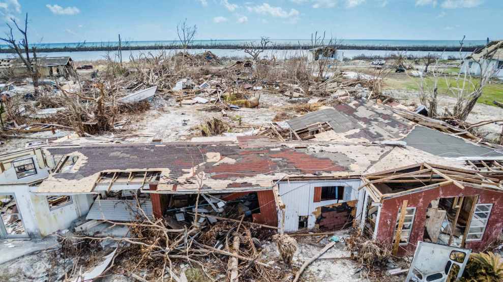 A destroyed home surrounded by broken trees with the ocean and blue skies in the background