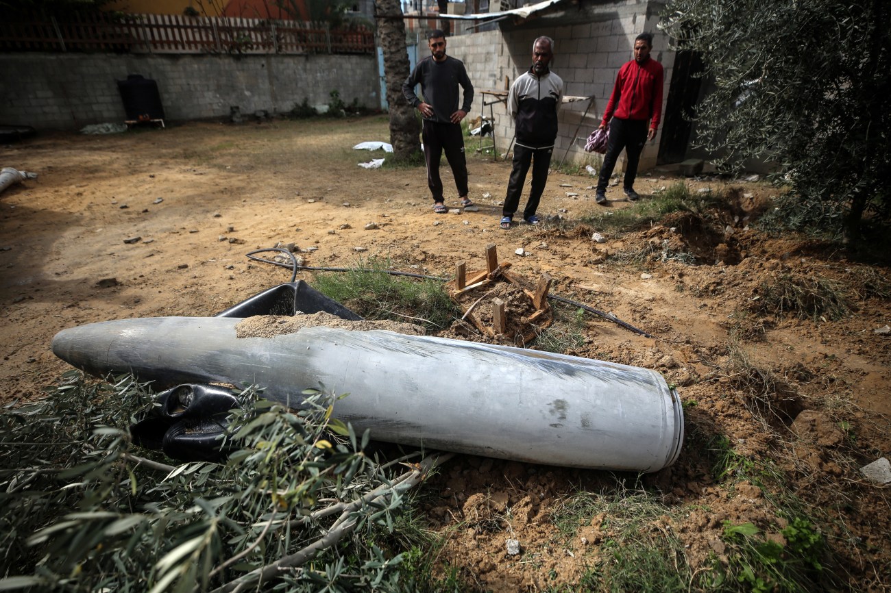 Three men stand next to unexploded bomb on the ground.