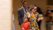 Arizona Senator Kyrsten Sinema looks at her watch as, in a blur, she walks past photographers in a yellow, pink and black dress while holding a red handbag.