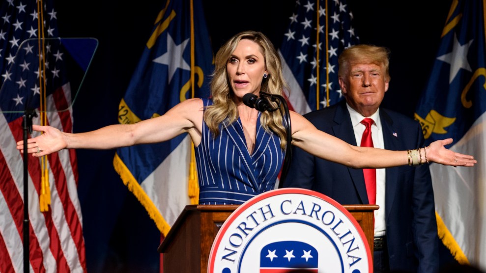 Lara Trump stands at a podium with her arms wide, speaking to a crowd in North Carolina while Donald Trump stands behind her.
