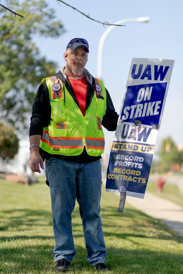 Full-length portrait of a man in a yellow vest holding UAW strike signs.
