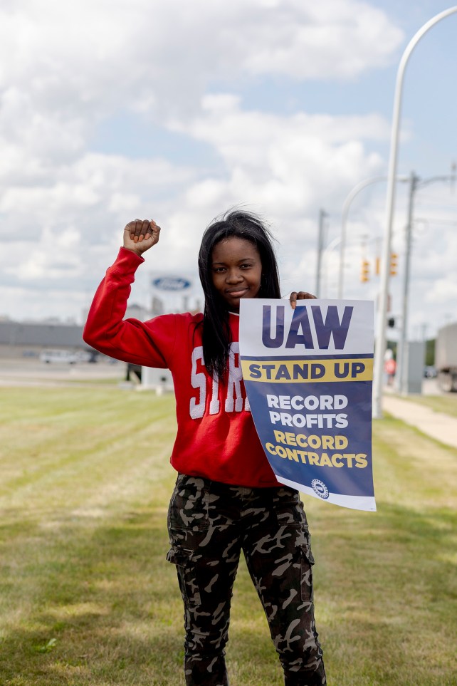 Young woman in red sweatshirt with her fist raised holding a UAW strike poster.